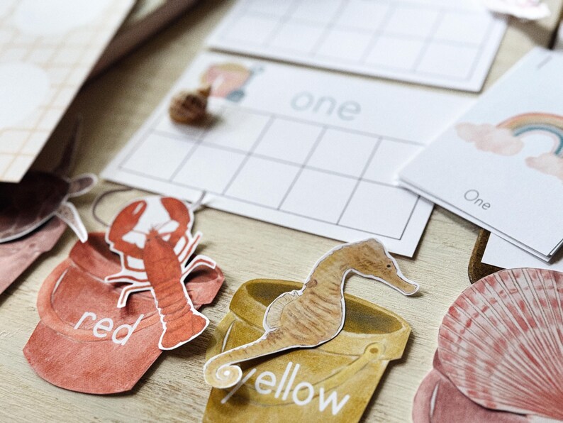 May include: A collection of watercolour illustrations and educational cards. Features a red lobster, a seahorse, and a seashell. The cards display the word "one" and a grid for counting. The colour words "red" and "yellow" are also present.