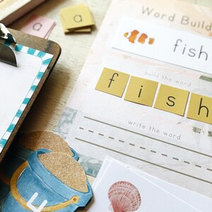 May include: Educational word-building set with the word "fish". Includes letter tiles, a worksheet with dotted lines for writing, and a clownfish illustration. A blue and yellow bucket is also visible.