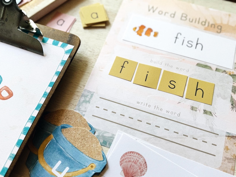 May include: Educational word-building set with the word "fish". Includes letter tiles, a worksheet with dotted lines for writing, and a clownfish illustration. A blue and yellow bucket is also visible.