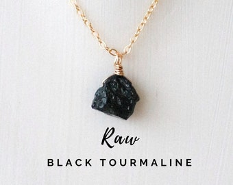 May include: Raw black tourmaline pendant necklace on a gold chain. The pendant is a rough, natural piece of black tourmaline.