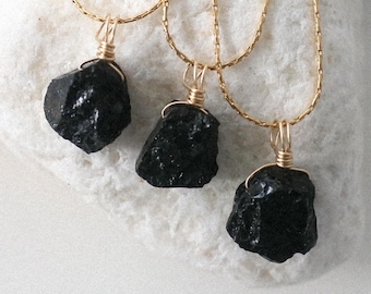 May include: Three black tourmaline necklaces with gold-colored chains and wire wrapping. The raw, natural gemstones are displayed on a white, textured surface, showcasing their dark, textured appearance. The necklaces are a type of jewelry.