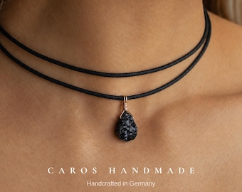 Puede incluir: Un collar de doble cordón negro con un colgante de piedra rugosa, oscura y texturizada, envuelto en un fino alambre metálico. El texto CAROS HANDMADE Handcrafted in Germany aparece impreso en blanco en la parte inferior.