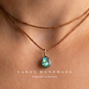 Könnte beinhalten: Eine doppelte braune Kordelkette mit einem rohen, schillernden blau-grünen Labradorit-Anhänger, der mit goldfarbenem Draht umwickelt ist. Der Text CAROS HANDMADE Handcrafted in Germany steht in Weiß am unteren Bildrand.