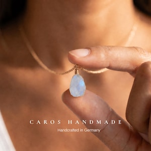 May include: A teardrop-shaped moonstone pendant suspended on a thin gold chain. The stone features a translucent, iridescent blue glow. The text CAROS HANDMADE Handcrafted in Germany is printed at the bottom of the image in white lettering.