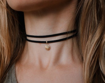 May include: A black double-layered choker necklace with a small gold pendant. The pendant features a sun design. The necklace is worn around the neck.