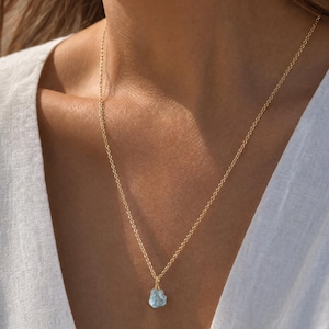 May include: A delicate gold chain necklace featuring a single, small, raw, light blue gemstone pendant. The stone has an irregular, natural shape and is securely wrapped in thin gold wire, suspended from a simple, fine-link gold metal chain.