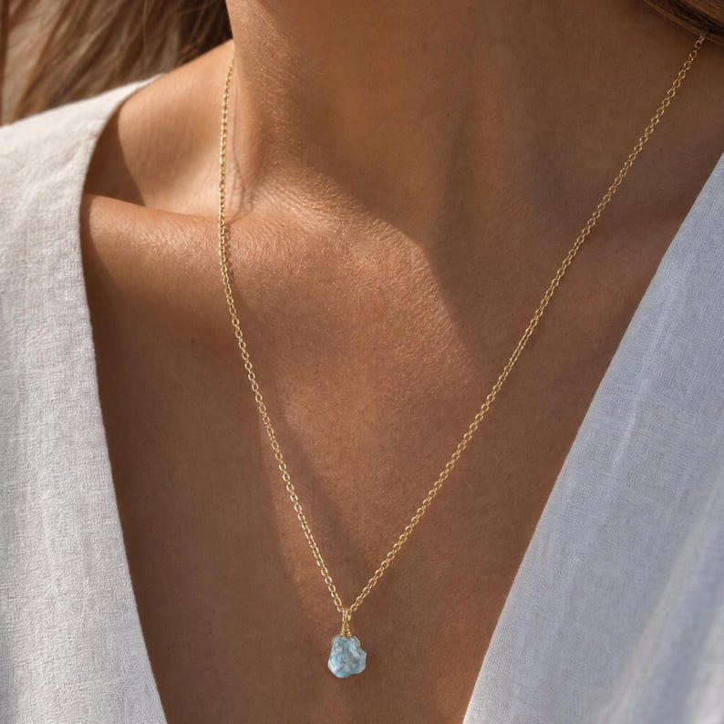 May include: A delicate gold chain necklace featuring a single, small, raw, light blue gemstone pendant. The stone has an irregular, natural shape and is securely wrapped in thin gold wire, suspended from a simple, fine-link gold metal chain.