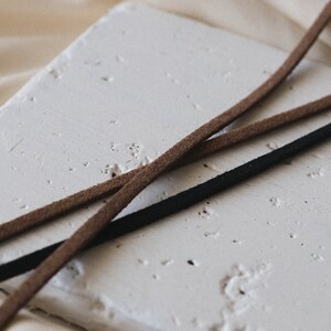 May include: Two leather cords, one brown and one black, are lying on a white textured surface. The cords are crossed over each other.