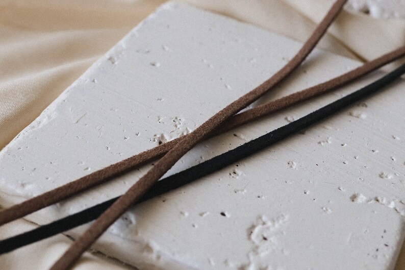 May include: Two leather cords, one brown and one black, are lying on a white textured surface. The cords are crossed over each other.