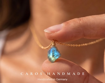 May include: A raw, iridescent blue and green labradorite gemstone pendant wrapped in gold-colored wire, suspended from a delicate gold-tone chain. The text CAROS HANDMADE Handcrafted in Germany is printed at the bottom of the image.