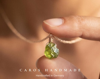 May include: A raw, translucent olive-green peridot gemstone pendant suspended on a delicate gold-tone chain. The stone is secured with a gold-colored wire wrap at the top. The text CAROS HANDMADE Handcrafted in Germany is visible at the bottom.