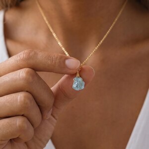 May include: A gold chain necklace featuring a single raw, light blue aquamarine gemstone pendant. The stone is secured with gold wire wrapping at the top, connecting it to the delicate gold link chain that rests around the neck area.