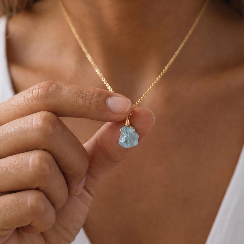 May include: A gold chain necklace featuring a single raw, light blue aquamarine gemstone pendant. The stone is secured with gold wire wrapping at the top, connecting it to the delicate gold link chain that rests around the neck area.