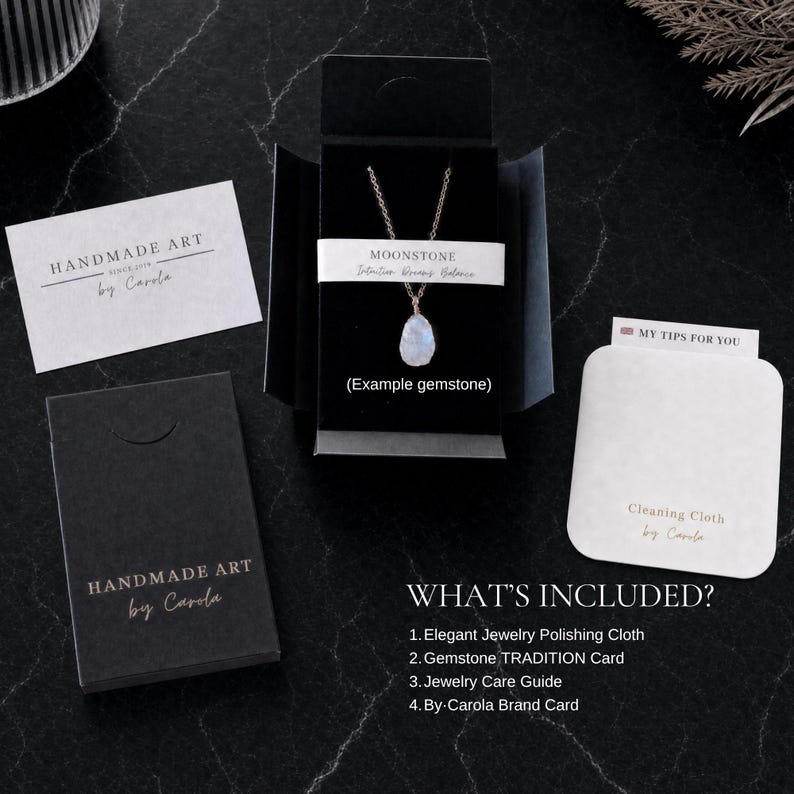 May include: A necklace with a teardrop-shaped moonstone pendant on a thin chain, presented in a black box. Includes a white card labeled HANDMADE ART by Carola, a cleaning cloth labeled MY TIPS FOR YOU, and a list detailing the included items.