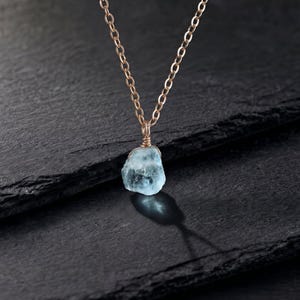 May include: A necklace featuring a raw, light blue gemstone pendant wrapped in thin gold-toned wire. The pendant hangs from a delicate gold-toned metal chain with oval links. The gemstone has a translucent, irregular, and textured appearance.