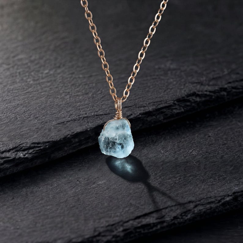 May include: A necklace featuring a raw, light blue gemstone pendant wrapped in thin gold-toned wire. The pendant hangs from a delicate gold-toned metal chain with oval links. The gemstone has a translucent, irregular, and textured appearance.