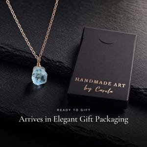 May include: A necklace featuring a raw, light blue gemstone pendant wrapped in gold-toned wire on a matching chain, positioned next to a black rectangular box with the gold-printed text HANDMADE ART by Carola, READY TO GIFT, Arrives in Elegant Gift Packaging.