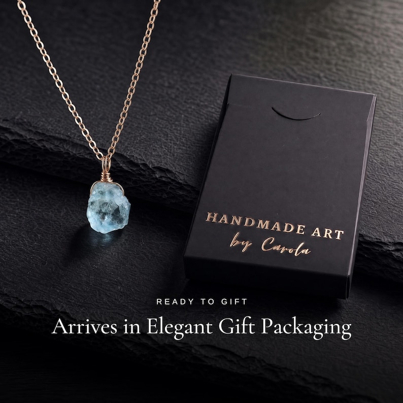 May include: A necklace featuring a raw, light blue gemstone pendant wrapped in gold-toned wire on a matching chain, positioned next to a black rectangular box with the gold-printed text HANDMADE ART by Carola, READY TO GIFT, Arrives in Elegant Gift Packaging.