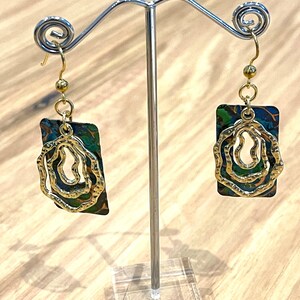 May include: A pair of gold and green earrings with a swirling design. The earrings are hanging from a silver stand.