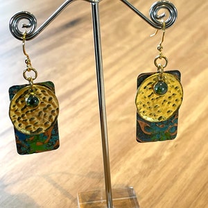 May include: A pair of gold and green earrings with a hammered metal design. The earrings are hanging from a silver stand.