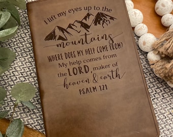 May include: A brown leather journal with the words "I lift my eyes up to the mountains" and "Where does my help come from?" engraved on the front. The text continues with "My help comes from the Lord, maker of heaven & earth" and "Psalm 121".