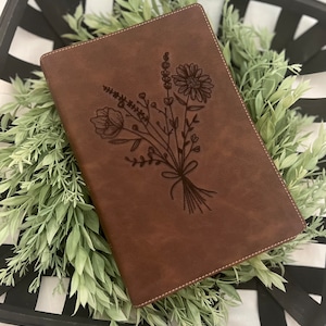 May include: A brown leather journal with a floral design embossed on the front cover. The journal is sitting on a black and white checkered surface.