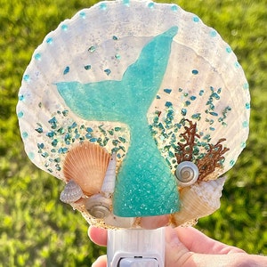 May include: A teal mermaid tail night light made of resin with a seashell design. The night light is shaped like a seashell and has a teal mermaid tail in the center. The seashell is decorated with glitter and small seashells.