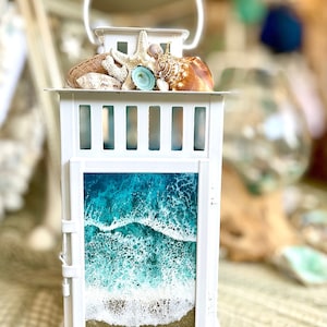 May include: White lantern with a seashell and starfish design on top. The lantern has a glass panel that shows a resin ocean scene with blue water and white waves.