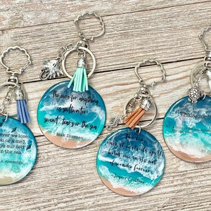 May include: Four round keychains with ocean-themed designs. Each keychain features a quote about the sea, with a blue and white wave design, a tassel, and a charm. The keychains have silver-colored hardware.
