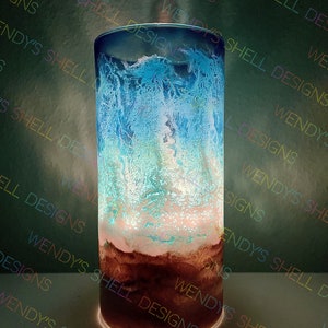 Puede incluir: Un cilindro de vidrio transparente con un dise&ntilde;o de resina azul, blanco y marr&oacute;n. La resina tiene un patr&oacute;n de remolino en forma de onda. El cilindro est&aacute; iluminado desde abajo, creando un brillo c&aacute;lido.