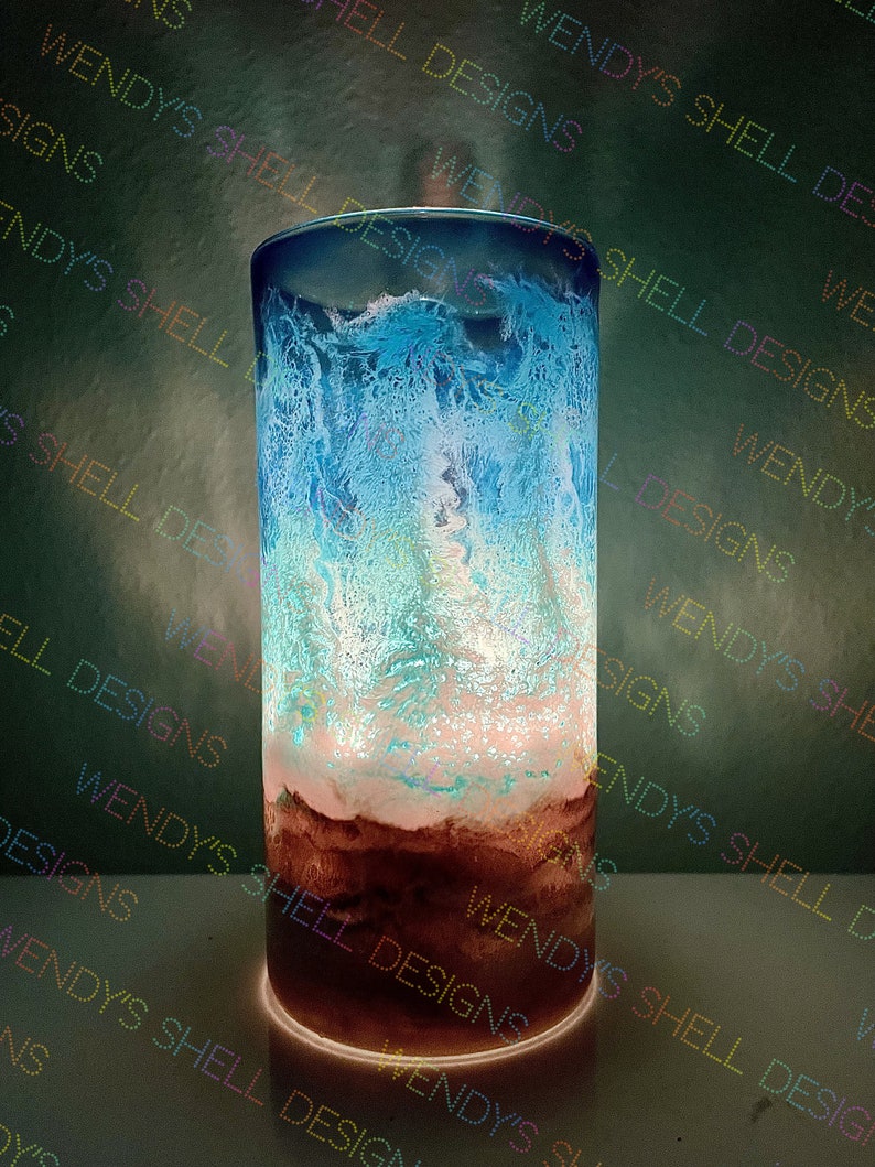 Puede incluir: Un cilindro de vidrio transparente con un dise&ntilde;o de resina azul, blanco y marr&oacute;n. La resina tiene un patr&oacute;n de remolino en forma de onda. El cilindro est&aacute; iluminado desde abajo, creando un brillo c&aacute;lido.