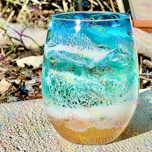 May include: A clear glass wine glass with a blue, white, and gold ocean-themed design. The glass is filled with resin that resembles waves crashing on a beach.