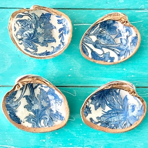 May include: Four decorative seashells with a blue and white floral pattern and gold trim. The shells are arranged in a square pattern on a turquoise wooden surface.