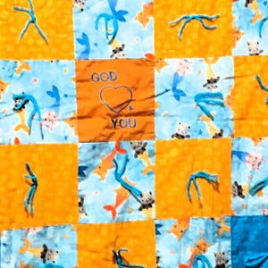 May include: A patchwork quilt with a blue and orange color scheme. The center square features the words "God" and "You" with a heart in between. The other squares feature a repeating pattern of fish and stars.