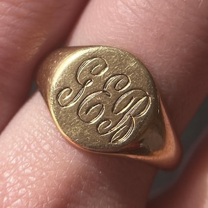 May include: A gold signet ring with a rounded face. The ring features an engraved monogram of the letters "GB" in an elegant script. The ring is a warm gold color and appears to be worn on a finger.