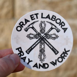 Op de afbeelding: Ronde witte sticker met zwarte tekst en een afbeelding van twee gekruiste hamers met een kruis in het midden. De tekst luidt "Ora et Labora Bid en werk".