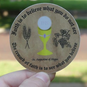 May include: A round sticker with a brown background and black text. The text reads "Faith is to believe what you do not see. The reward of faith is to see what you believe."  -St. Augustine of Hippo. The sticker also features a chalice with a host, wheat, and grapes.