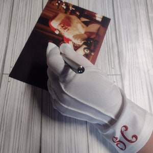 May include: White satin gloves with a red embroidered initial "S" on the cuff. The gloves are being worn by a hand holding a silver pen.