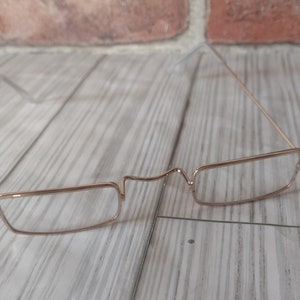 May include: A pair of antique-style, gold-colored eyeglasses with rectangular lenses. The glasses have a delicate, wire-frame design and a bridge that curves slightly. The background features a brick wall and a wooden surface.
