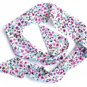 May include: A white silk scarf with a floral pattern in shades of pink, blue, and purple. The scarf is tied in a knot and is lying flat on a white background.