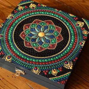 Puede incluir: Una caja de madera con un diseño de mandala pintado en la tapa. El mandala está hecho de pequeños puntos en tonos de verde, rojo, azul y dorado. La caja tiene un cierre de latón.