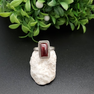 May include: A silver ring with a rectangular red gemstone set in a silver bezel. The ring is on a white rock.