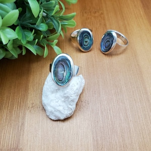 May include: Three silver rings with oval-shaped abalone shell inlays. The rings are on a wooden surface with a small white rock.