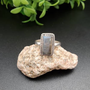 以下が含まれることがあります： 銀のリングに、長方形のラブラドライトの宝石が銀のベゼルにセットされています。リングは小さな岩の上に置かれています。
