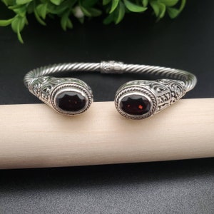 May include: A silver bangle bracelet with a twisted design and two red garnet stones set in the ends. The bracelet has an ornate design.