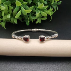 May include: A silver bangle bracelet with a twisted design and two square red gemstones. The bracelet has gold accents.