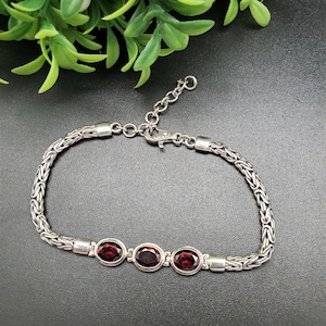 May include: A silver bracelet with three oval, deep red gemstones. The bracelet has a woven chain design and an adjustable clasp. The background is black with green foliage in the upper left corner.