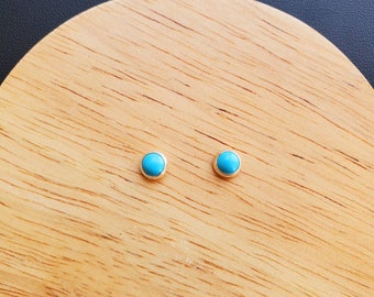 May include: A pair of small, round turquoise stud earrings with gold settings. The earrings are displayed on a wooden surface.