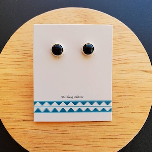 May include: A pair of black onyx stud earrings on a white card with the text "Sterling Silver" and a turquoise geometric border. The earrings have a sunburst design around the black stone.