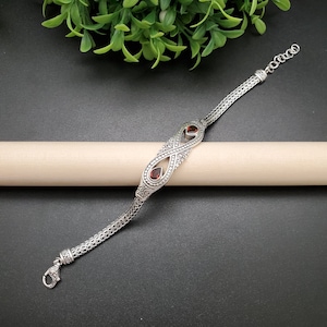 May include: A silver bracelet with an infinity symbol design featuring two red gemstones. The bracelet has a delicate chain link design.